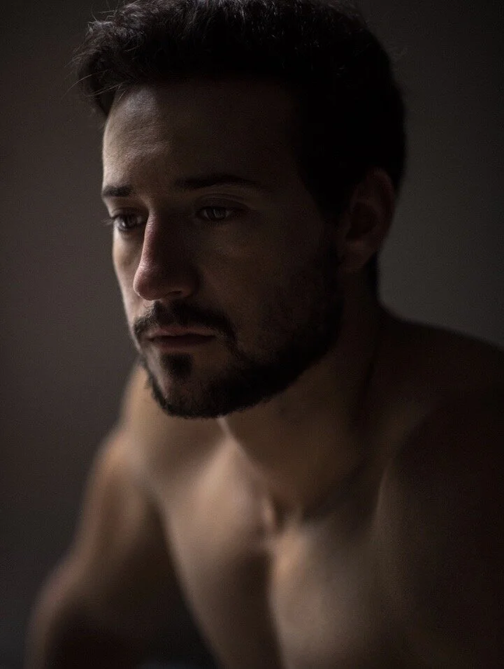 A shirtless man with a beard and short dark hair looking down in a dimly lit room.