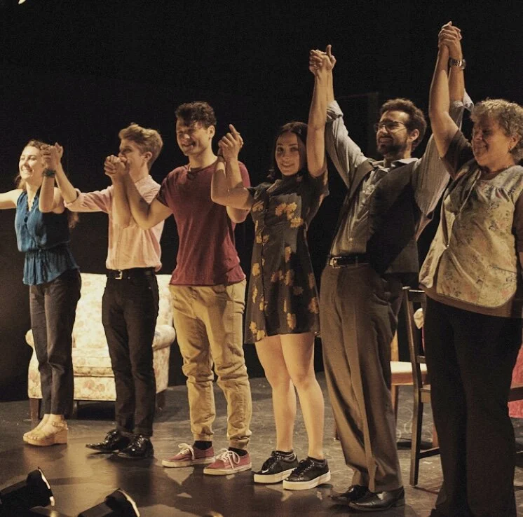 Group of six people standing on stage, holding hands and raising their arms in celebration after a performance.