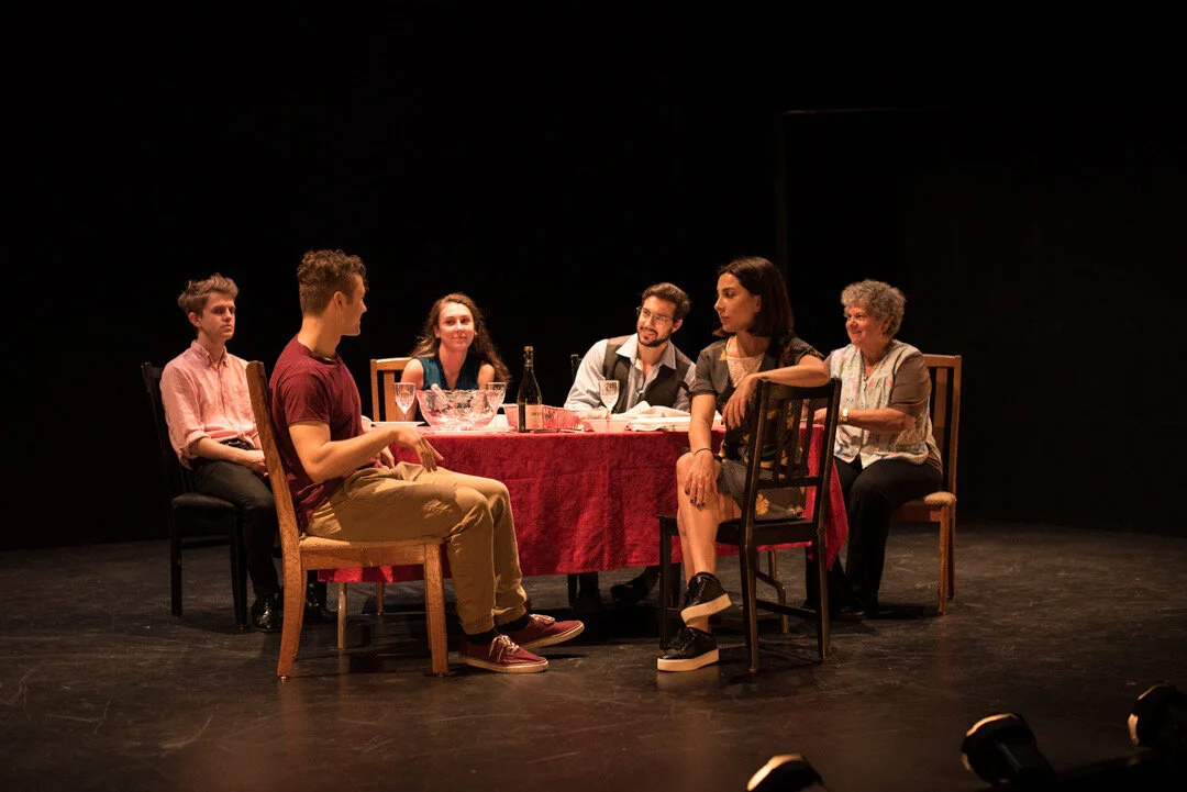 Group of six people sitting around a table with a red tablecloth on a stage, engaged in conversation during a theatrical performance.