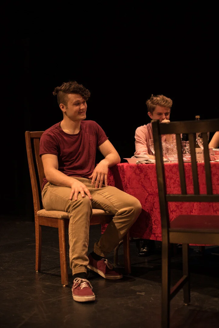 Two young men sitting at a table with a red tablecloth, one facing forward and the other partially obscured behind a chair, in a dimly lit setting, possibly on stage during a performance or dinner event.