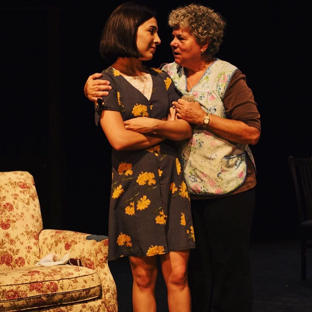Two women are standing close together, engaged in a serious conversation. The younger woman has shoulder-length dark hair and is wearing a dark dress with yellow floral patterns. The older woman has curly gray hair, is wearing a floral apron over a b