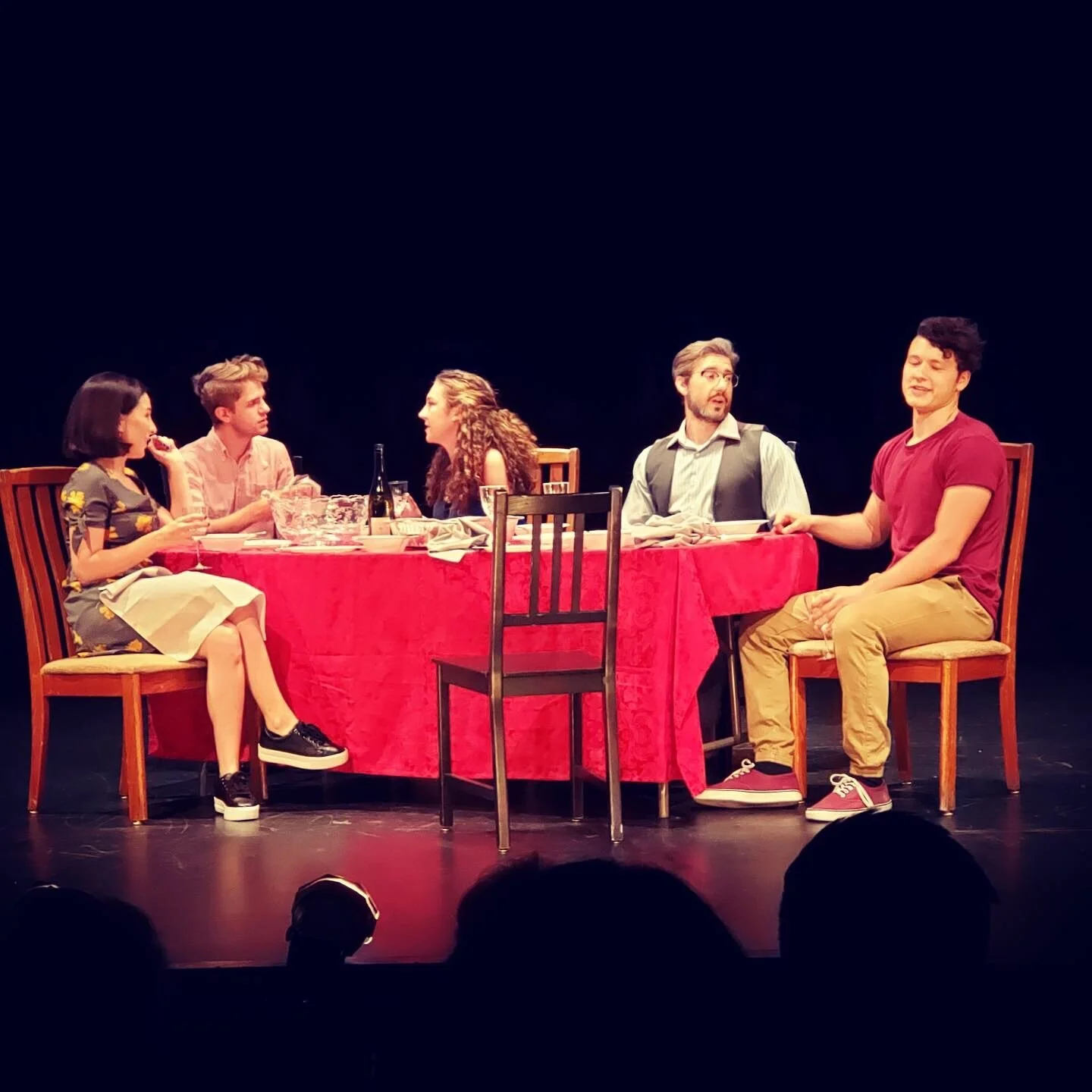 Six actors sitting around a red-draped dinner table, performing in a play, with a dark background.