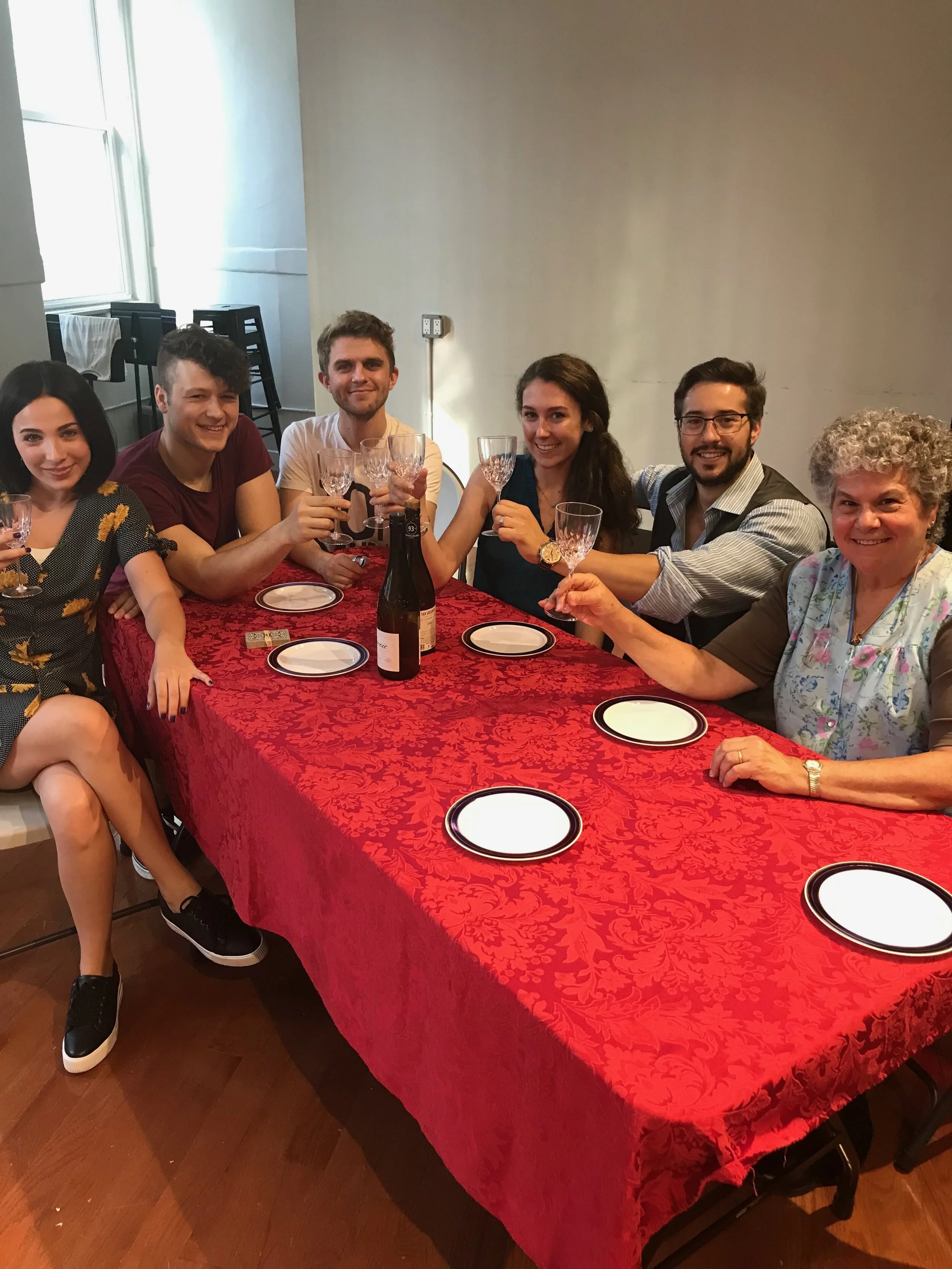 A group of six friends sitting at a table celebrating, raising glasses in a toast, with empty plates and a bottle of champagne on the table. The table is covered with a red tablecloth, and they are smiling at the camera.
