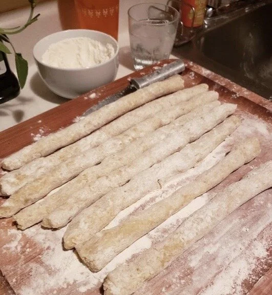 Unrolled dough strips dusted with flour on a wooden surface, with a bowl of flour, a glass of water, and a rolling pin in the background.