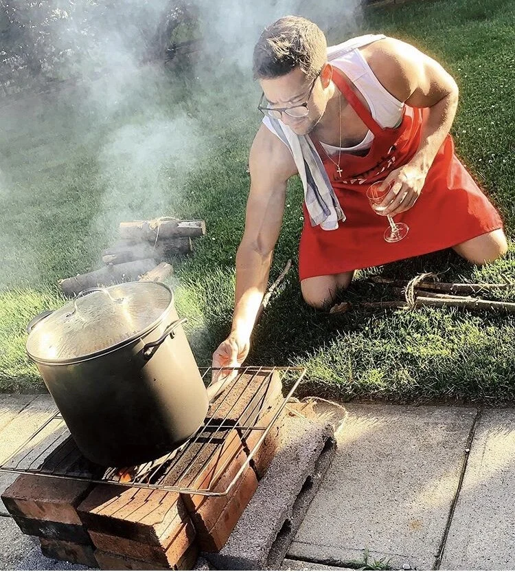 A man in a red apron and cross necklace kneeling on grass, holding a glass of wine, tending a large pot on an outdoor brick grill.
