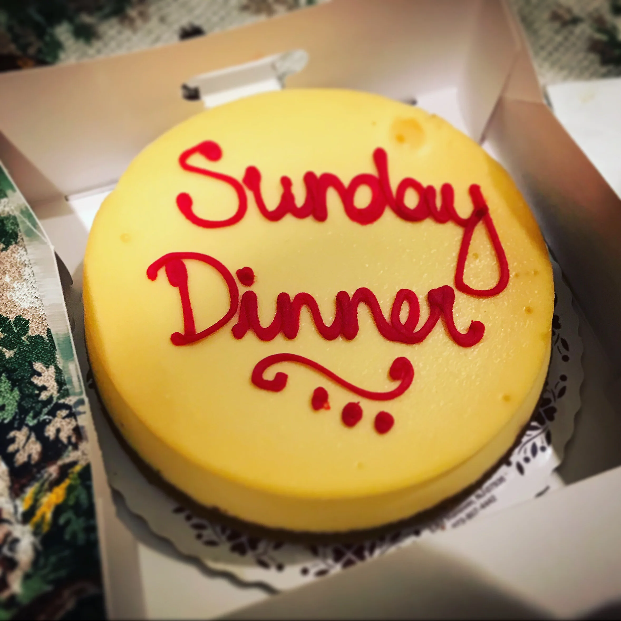 A round yellow cake with red icing writing on top inside a white cardboard box with the flaps open.