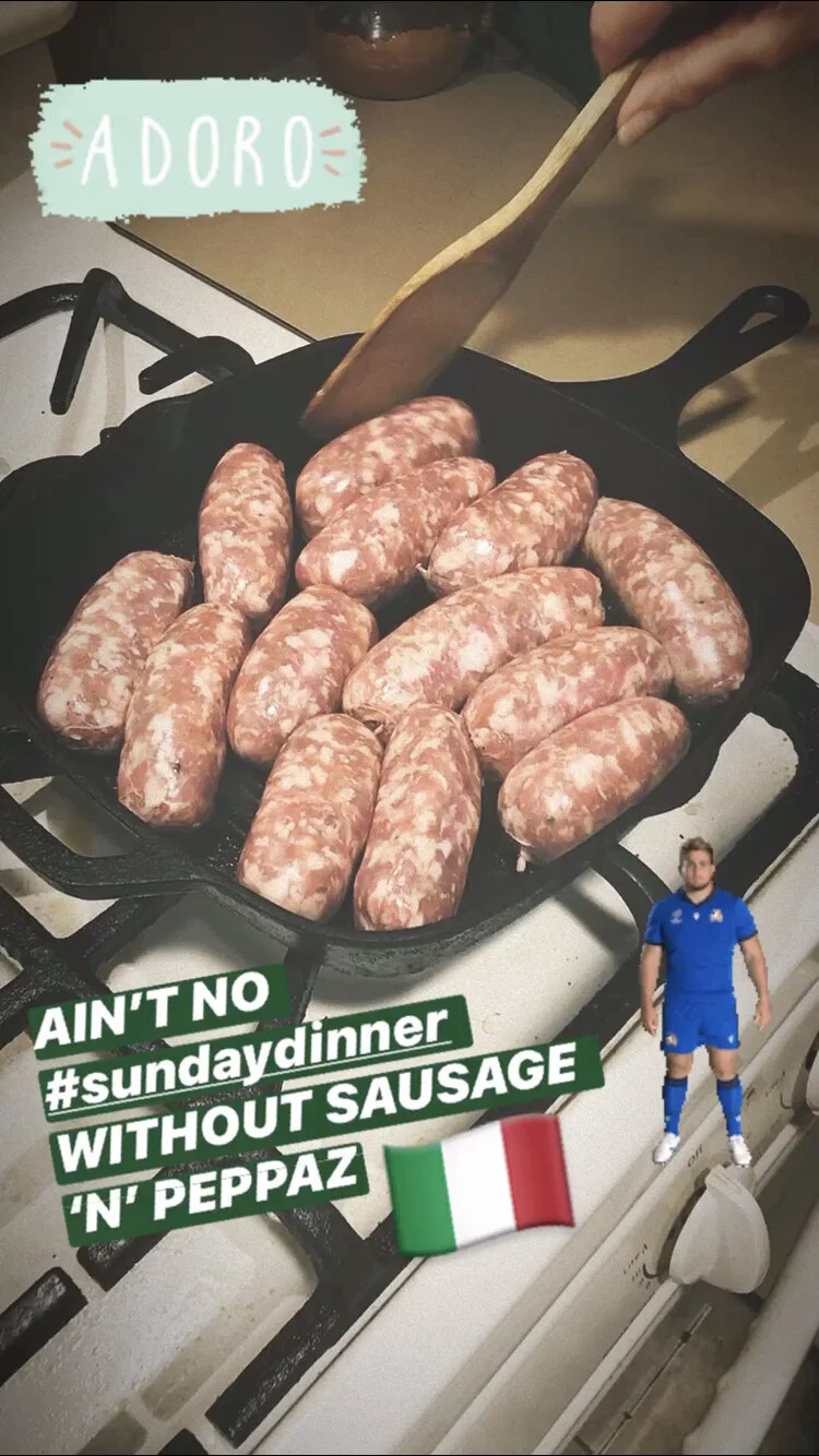 Raw sausages being cooked in a frying pan on a stove, with a hand holding a wooden spatula stirring them. There is a sticker with the word 'ADORO' and setup text that reads 'AIN'T NO #sundaydinner WITHOUT SAUSAGE 'N' PEPPADZ', along with a small Ital