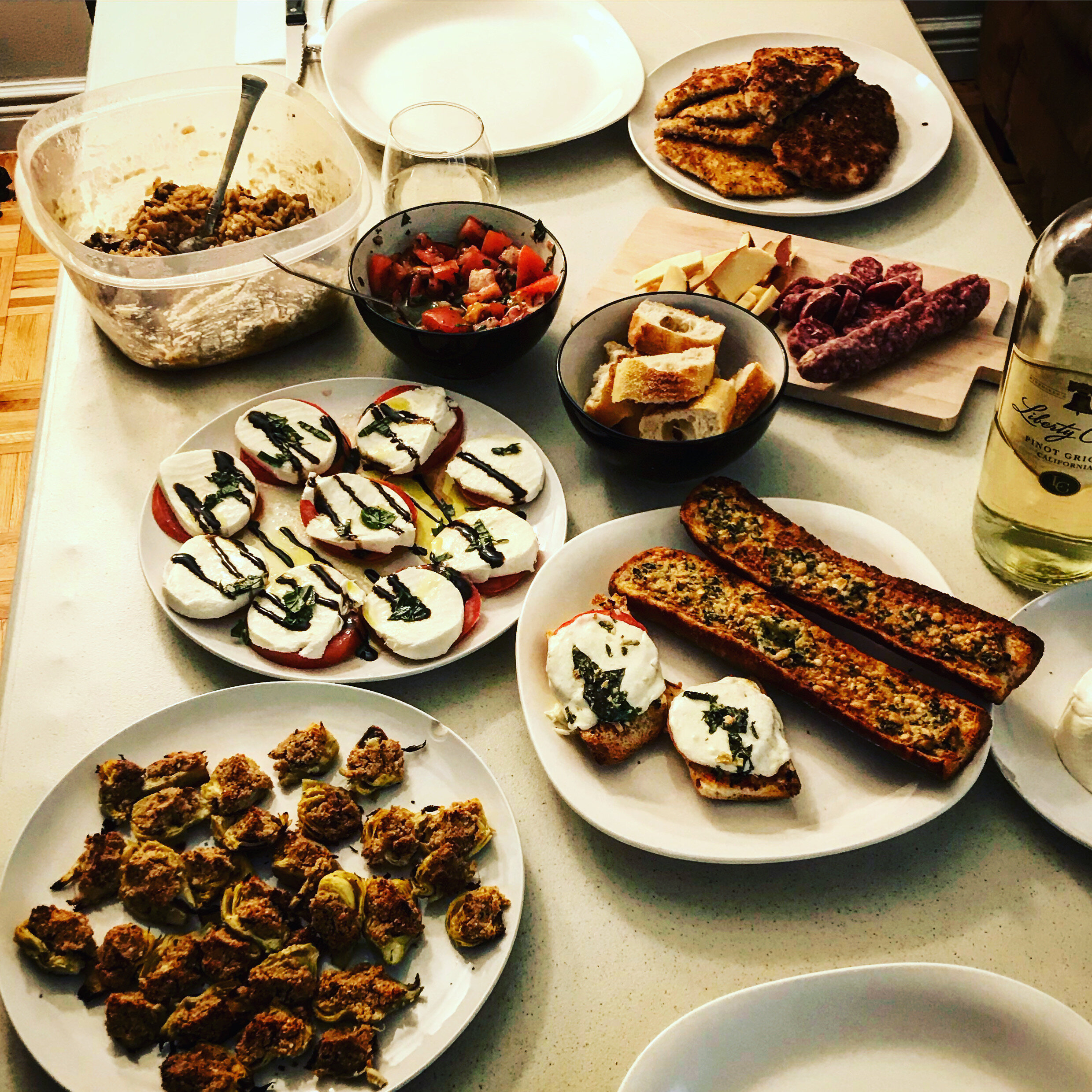Various plates and bowls of food on a kitchen counter, including Caprese salads, stuffed peppers, grilled bread, sliced tomatoes, cheese, and cooked sausage links.