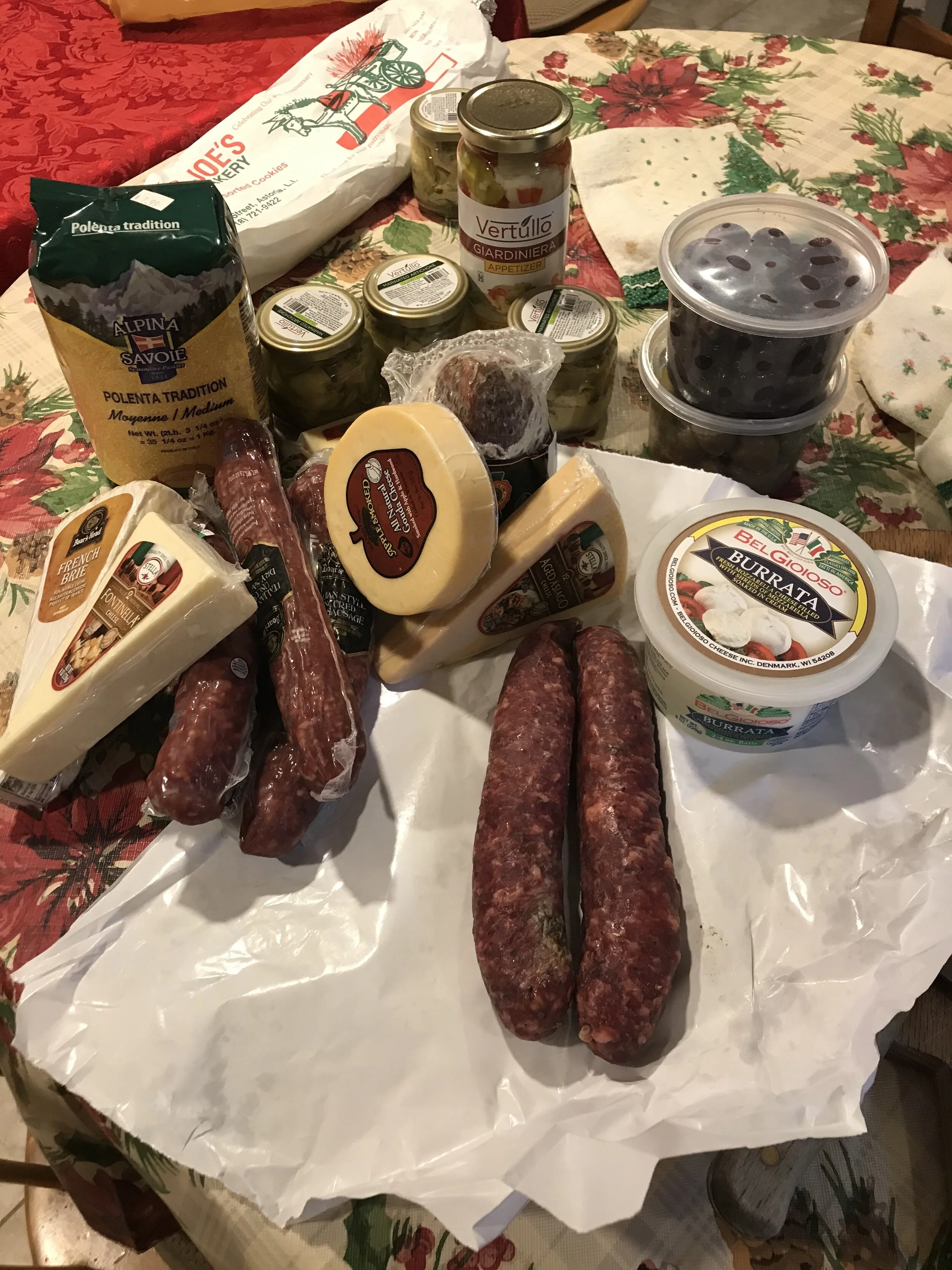 Freshly made Italian sausage placed on a table covered with parchment paper. The sausages are uncooked and appear to be finely ground with a red-seasoned exterior. There are various cheeses and other ingredients nearby, and the background has festive