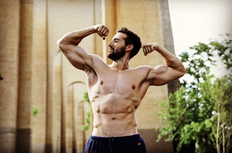 A muscular man is flexing his biceps outdoors in front of a building and some greenery.