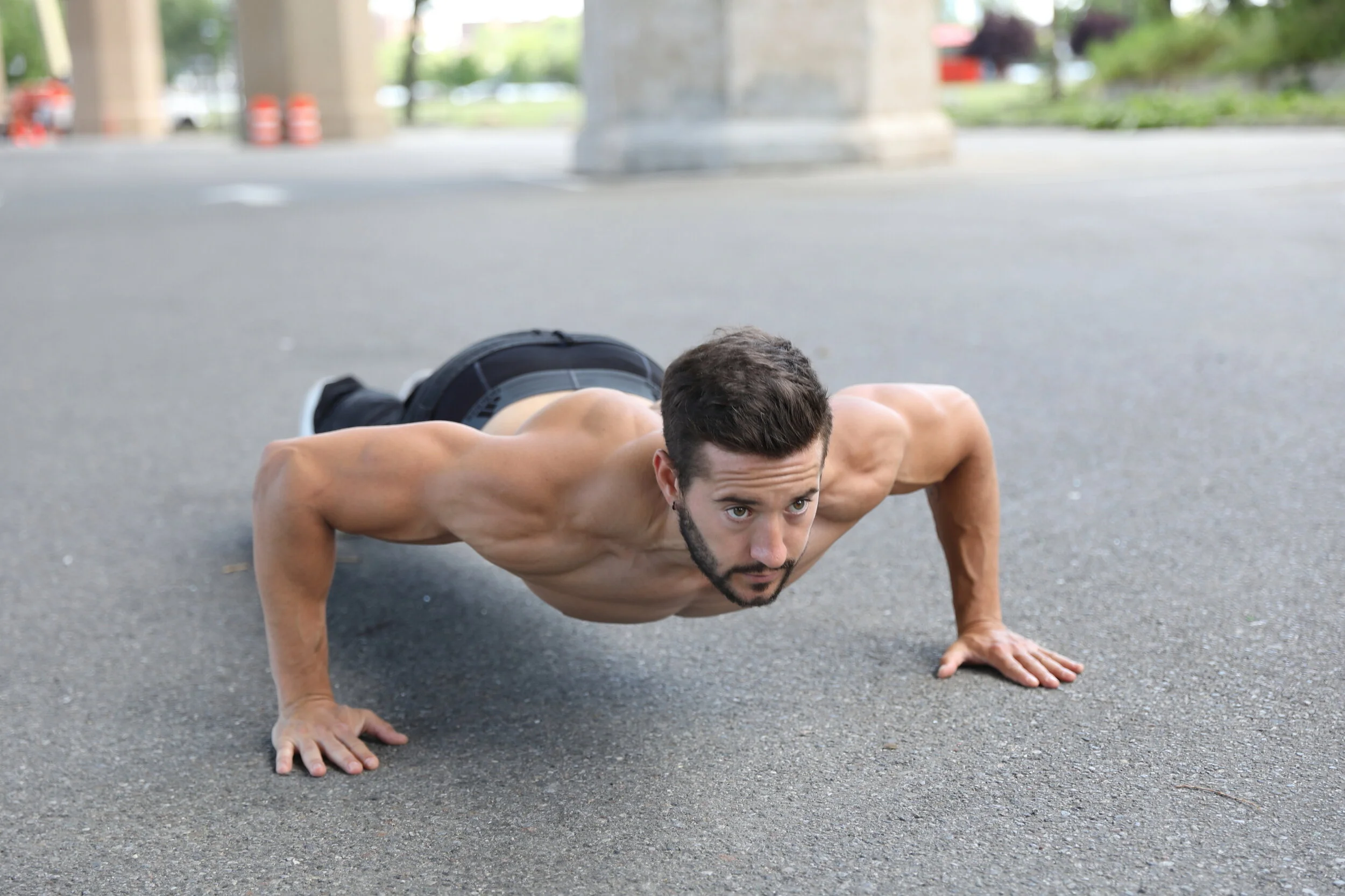 A man performing a push-up outdoors on a paved surface under a bridge or structure with concrete pillars.