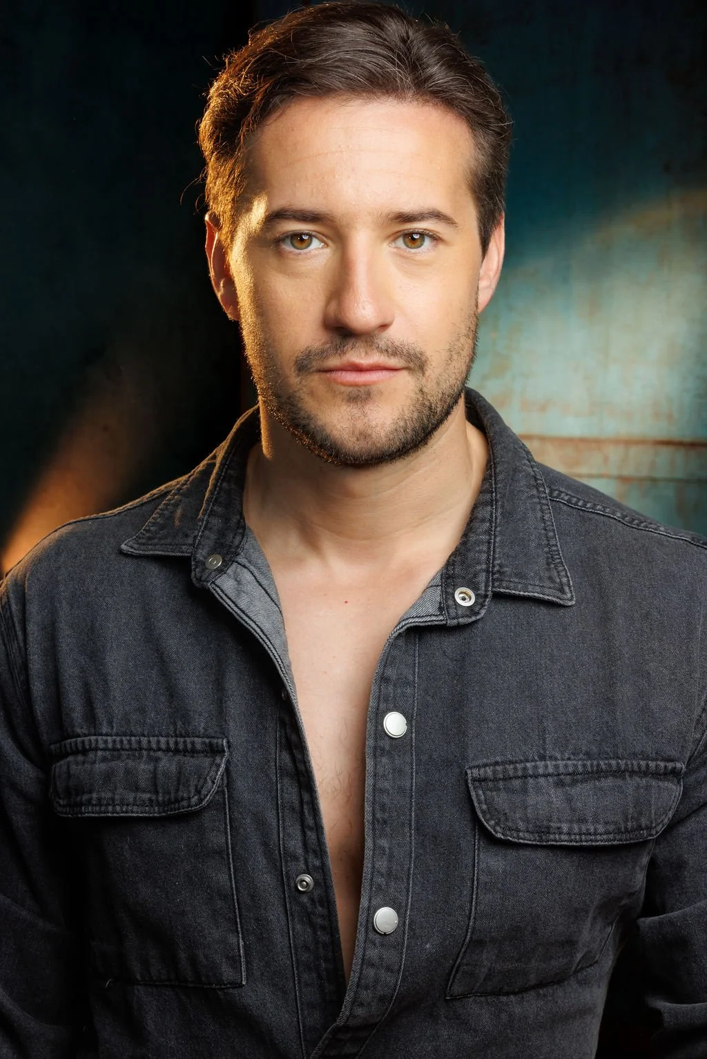 A man with brown hair, hazel eyes, and light facial hair, wearing a dark denim shirt, looking directly at the camera with a neutral expression, against a dark background with warm lighting.