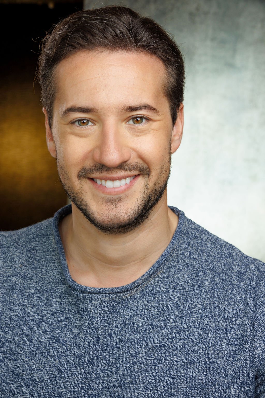 Head and shoulders portrait of smiling young man with brown hair, beard, wearing a blue sweater, holding camera in front of a blurred background.