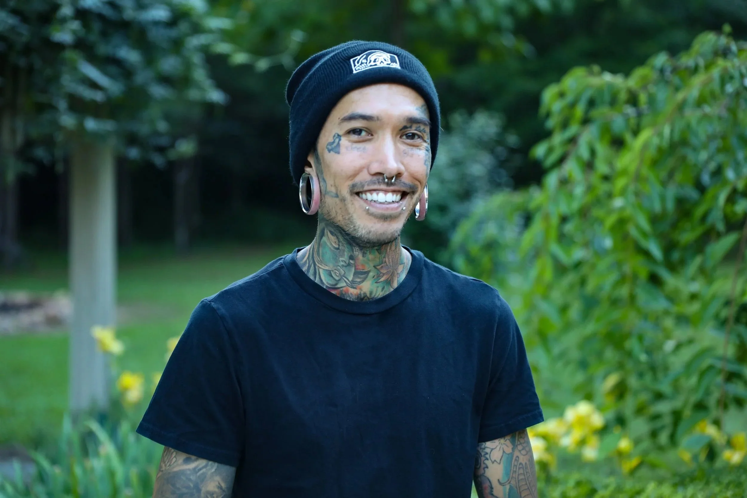 A smiling Shaun with tattoos, pierced ears, and facial tattoos wearing a black beanie and black t-shirt outdoors with green trees and plants in the background.