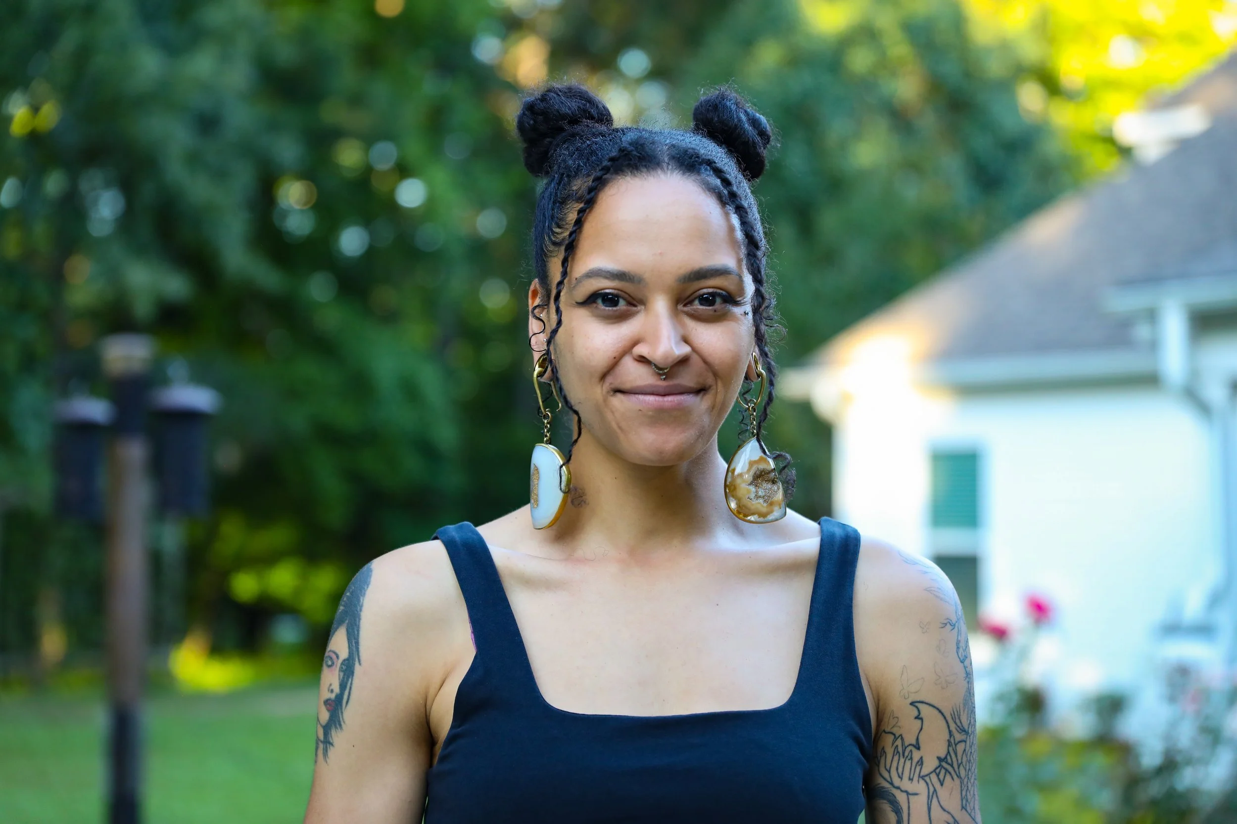 A Kat with dark hair styled in two buns and braids, wearing large earrings and a dark sleeveless top, standing outdoors with trees and a house in the background during daytime.