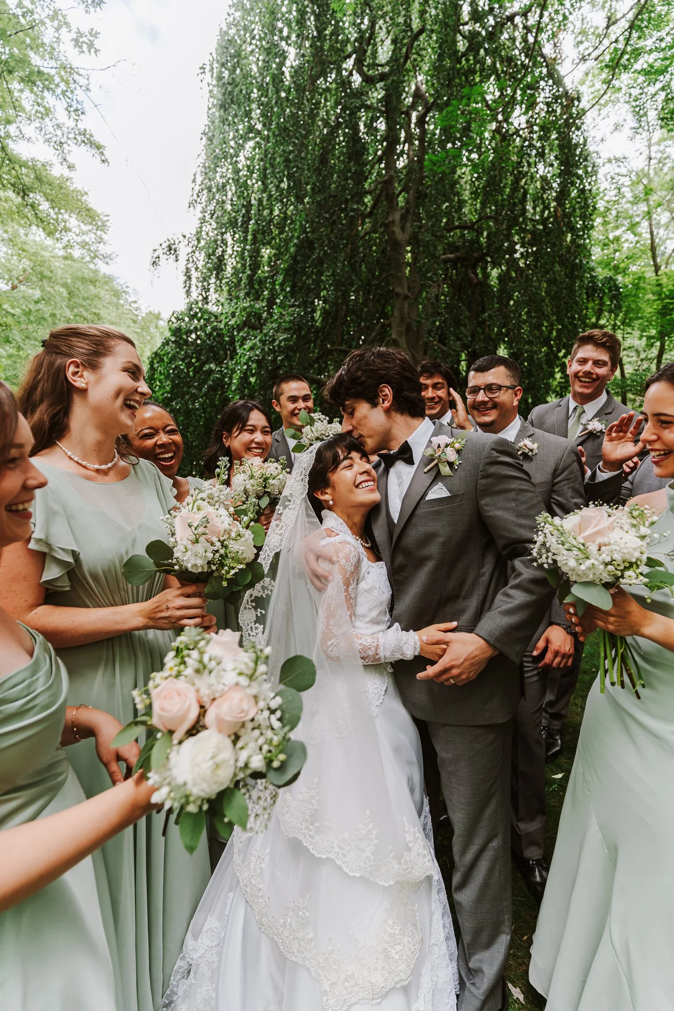 A Catholic wedding party gathered outdoors under trees, with the bride and groom in the center sharing a joyful moment, surrounded by bridesmaids holding bouquets, all smiling and celebrating.