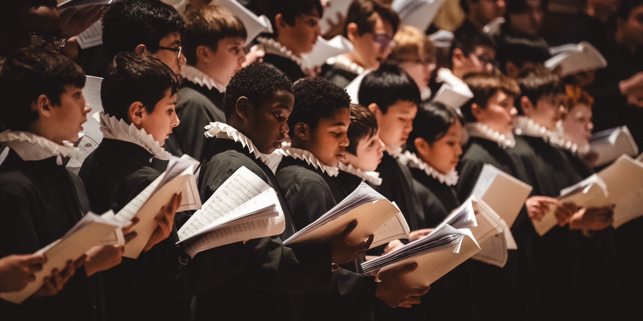 A Catholic choir of young boys dressed in black robes with white ruffled collars, holding sheet music and singing.