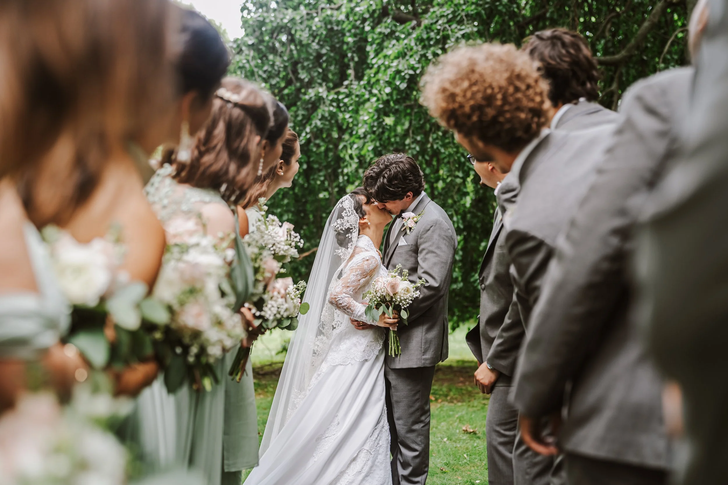 A Catholic bride and groom share a kiss at their outdoor wedding ceremony, with bridesmaids and groomsmen standing on either side in a lush green park setting.