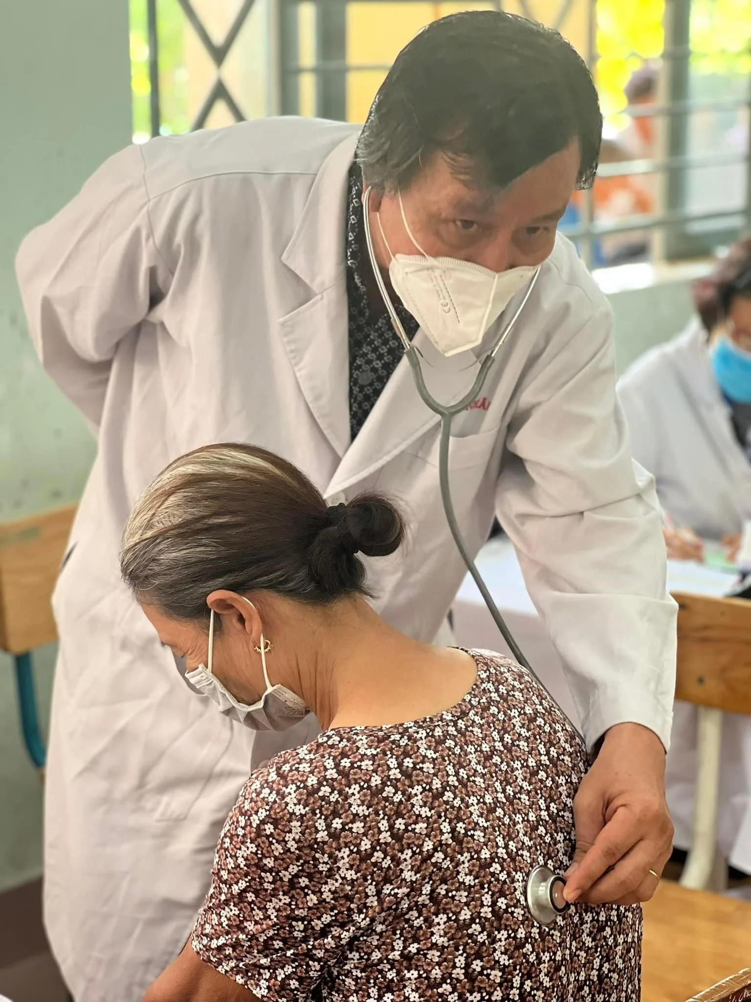 Masked doctor using a stethoscope to examine a seated woman in a clinical setting