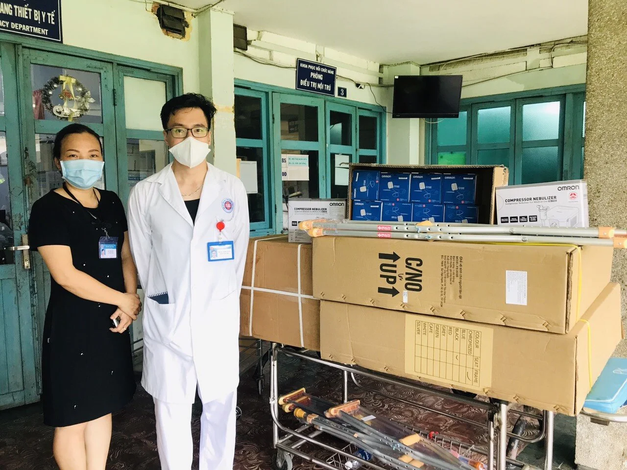 Masked healthcare workers stand beside boxed Omron compressor nebulizers and medical equipment outside a Vietnamese hospital ward.