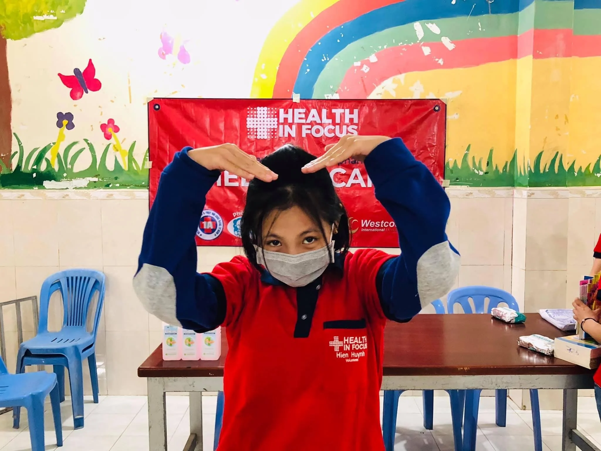 Masked Health in Focus volunteer forming a heart shape with her hands in front of a colorful rainbow mural