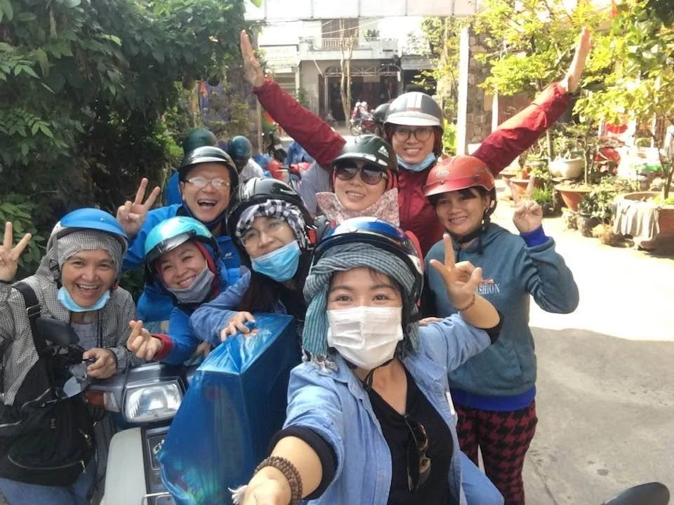 Group of helmeted women posing for a selfie on scooters in a sunny Vietnamese street