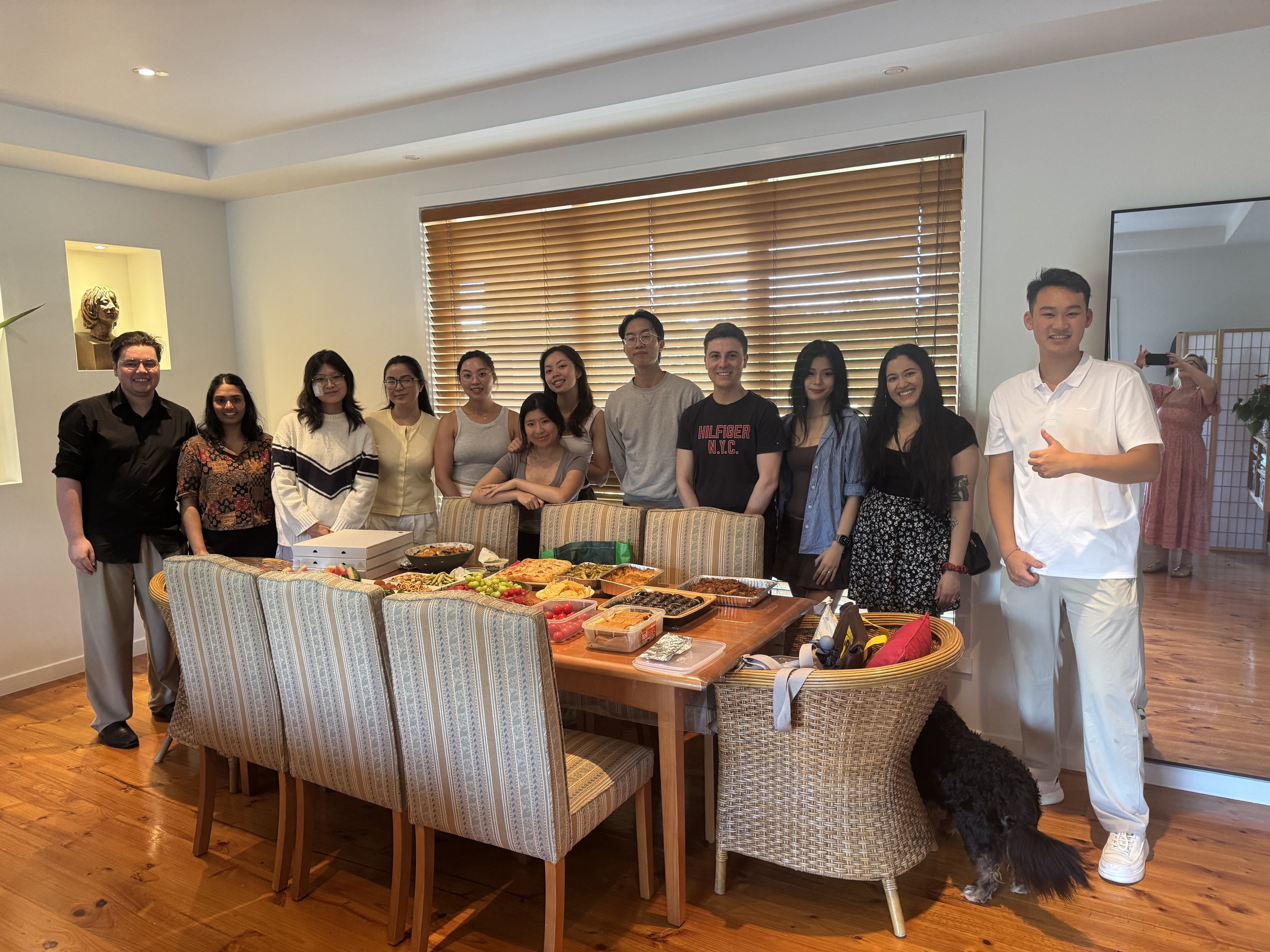 Group of people posing around a dining table filled with party food in a home, with a small black dog nearby