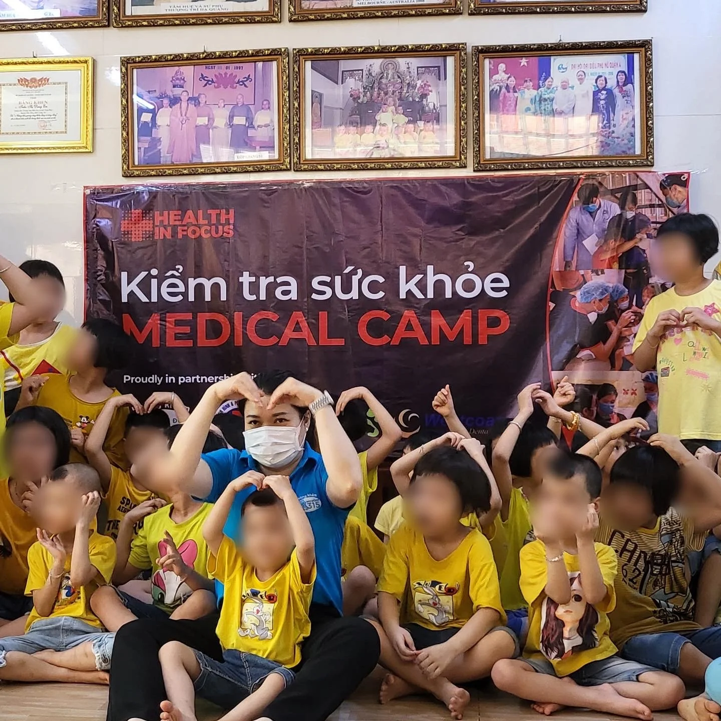 Children and volunteers pose in front of a Vietnamese medical camp banner, making heart shapes with their hands.