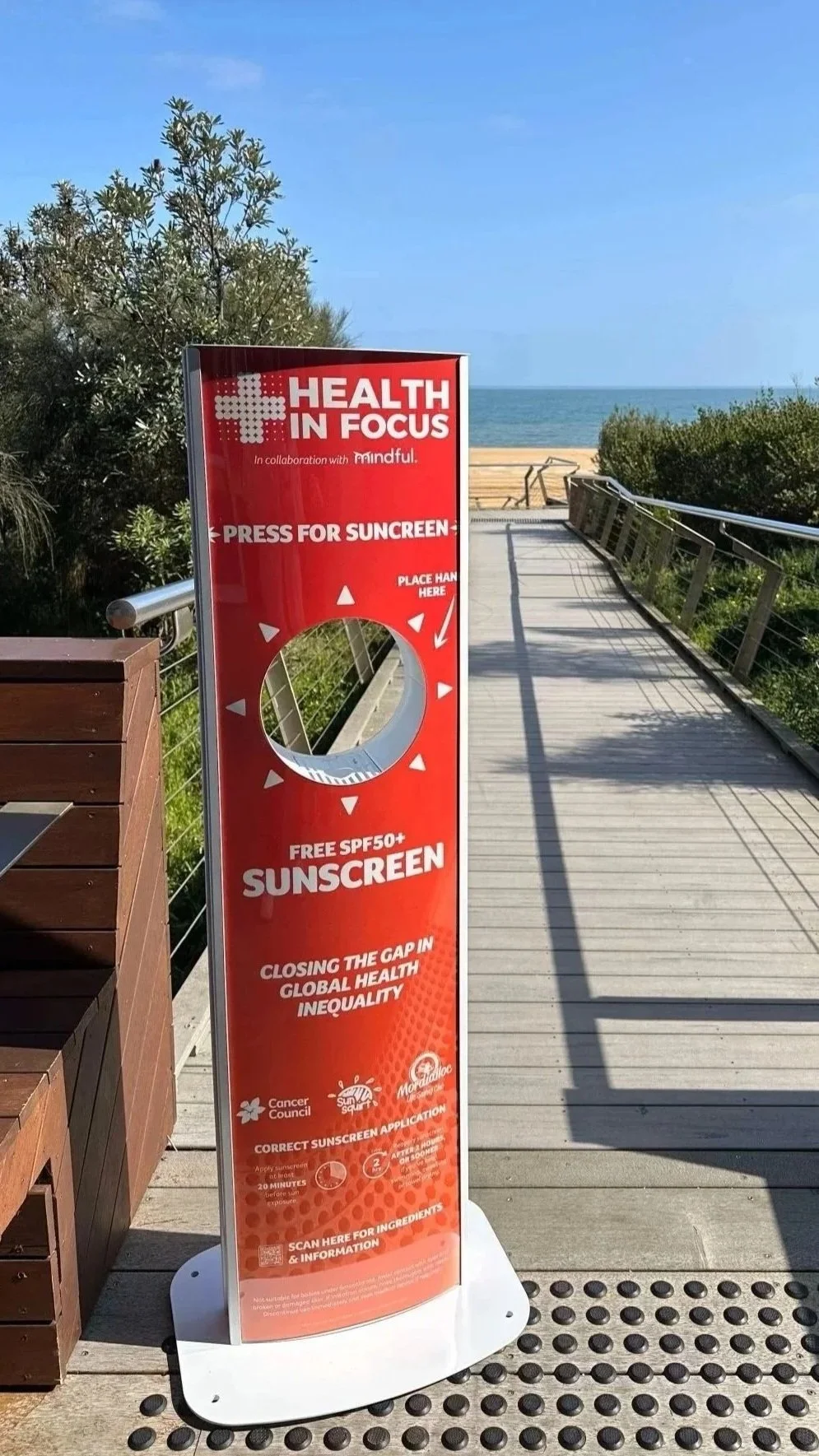 Red Health in Focus sunscreen sign offering free SPF 50+ sunscreen on a beach boardwalk overlooking the ocean