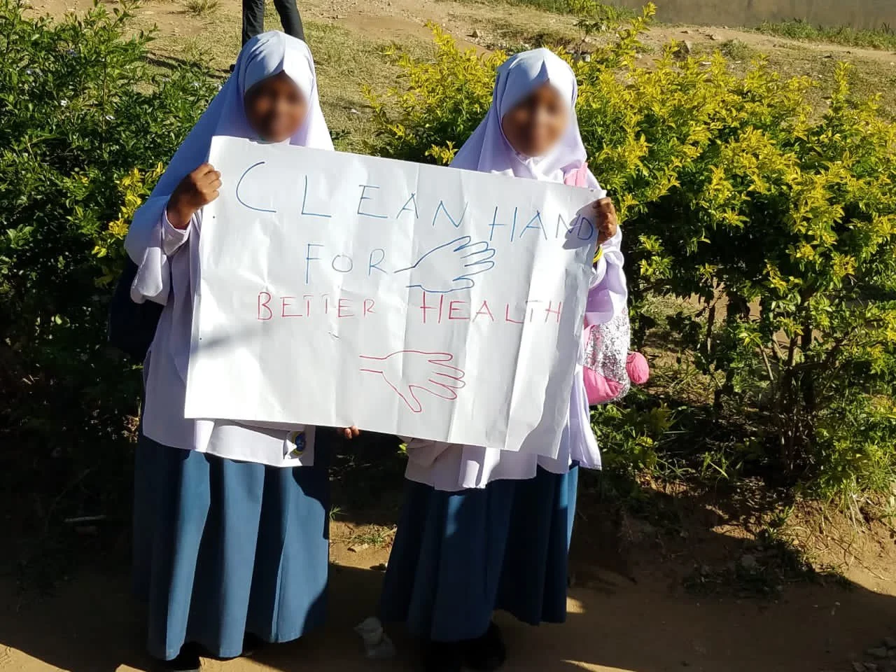 Two girls in hijabs hold a handmade sign reading “Clean hands for better health” outdoors.