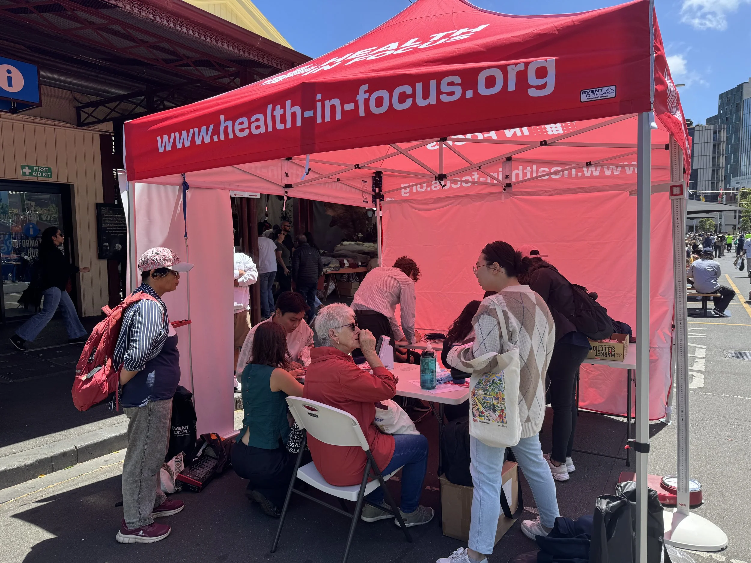 People gather at a health-in-focus community health screening tent on a sunny city street.