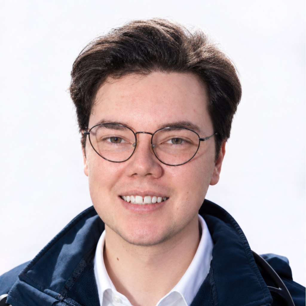 Smiling person wearing round glasses, a white shirt, and a dark jacket against a white background