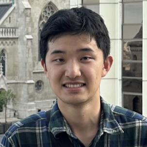 Portrait of a smiling young man wearing a green plaid shirt in front of a stone Gothic building
