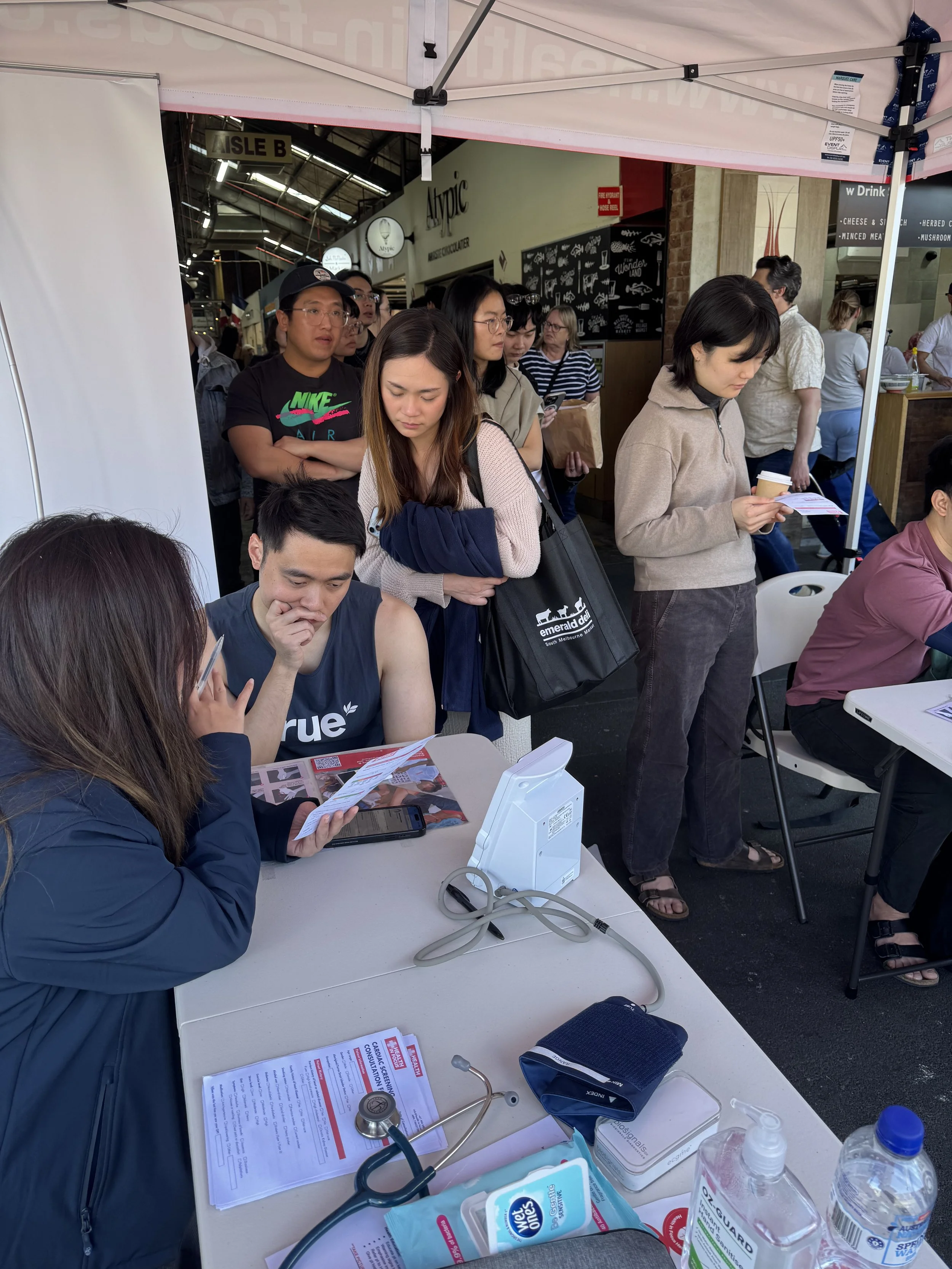 Crowded health screening booth at a market, with people waiting beside a table displaying a stethoscope, blood pressure cuff, medical forms, wipes, and water bottles.