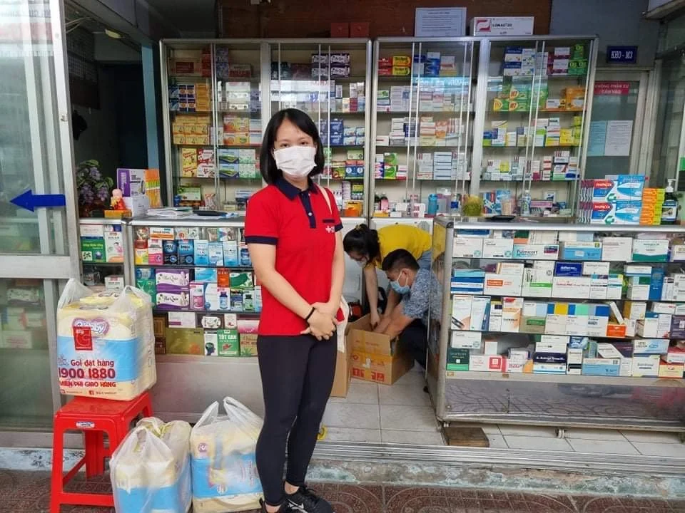 Masked pharmacy worker standing outside a pharmacy stocked with medicines, while two employees organize boxes inside