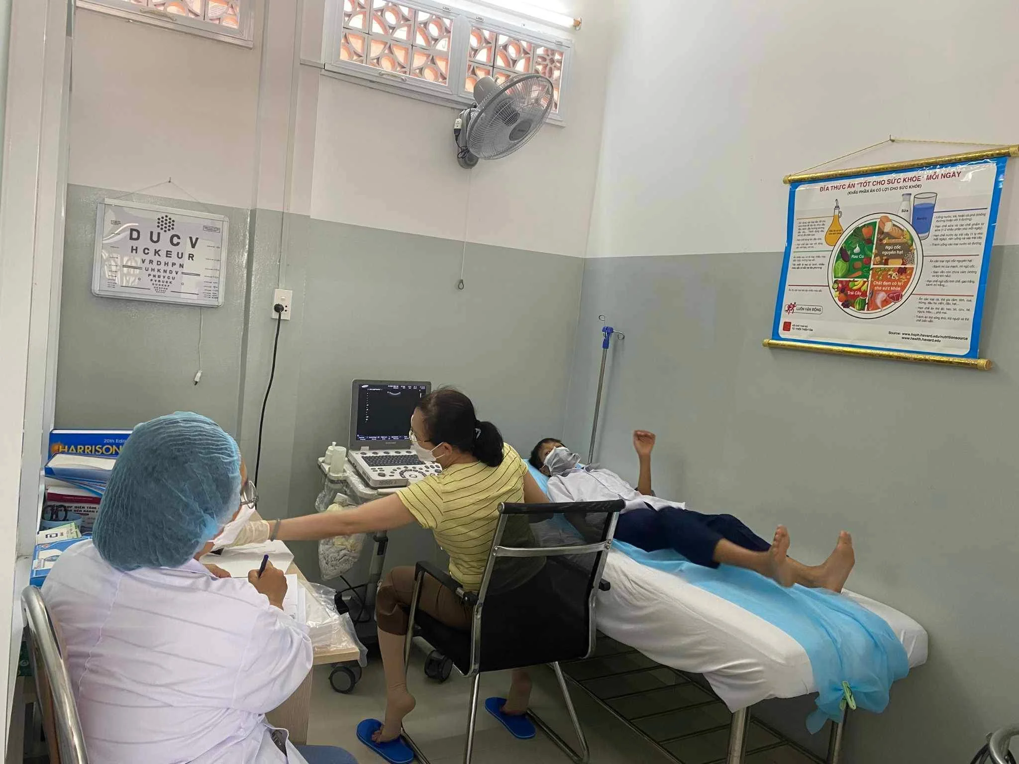 Medical clinic room with a masked patient lying on an examination bed while healthcare workers operate an ultrasound machine; Vietnamese health poster on the wall.