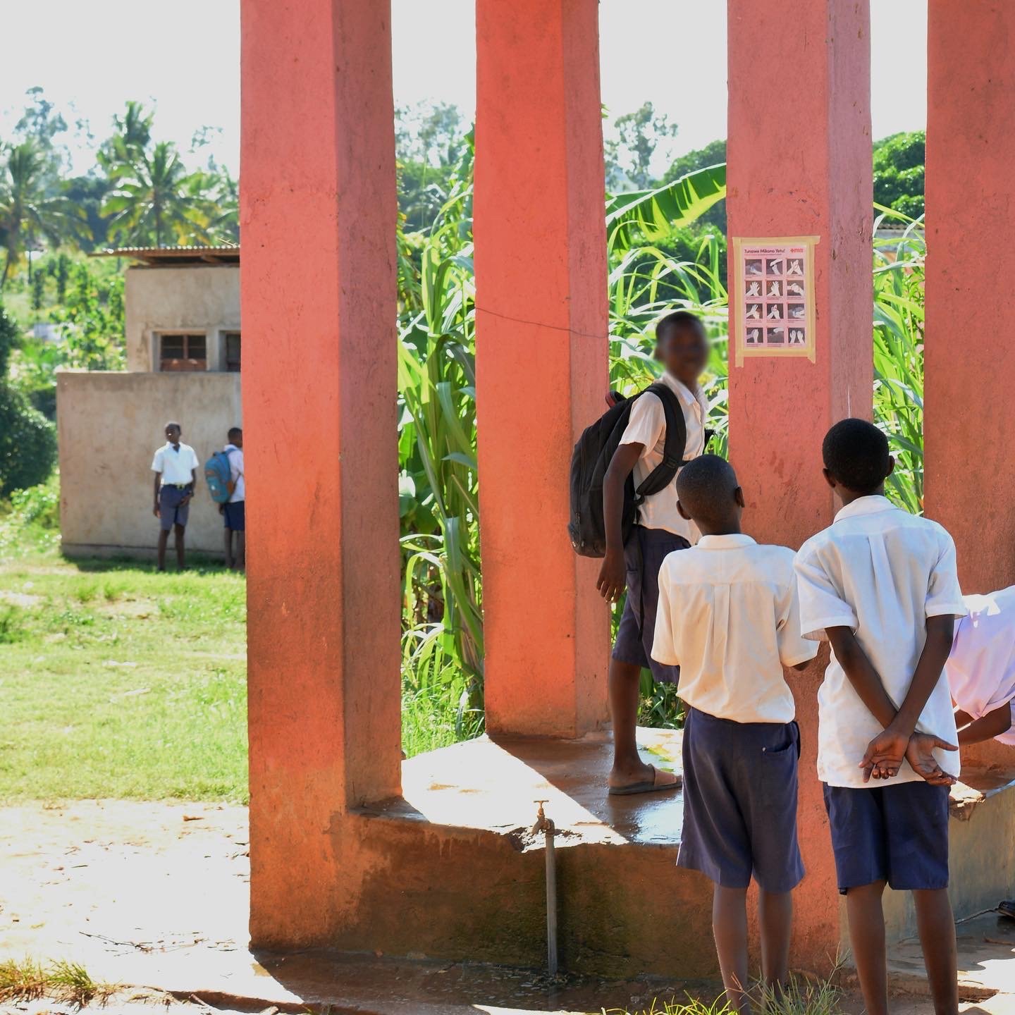 Schoolchildren in white shirts and blue shorts gather beside a water faucet under a covered outdoor walkway.