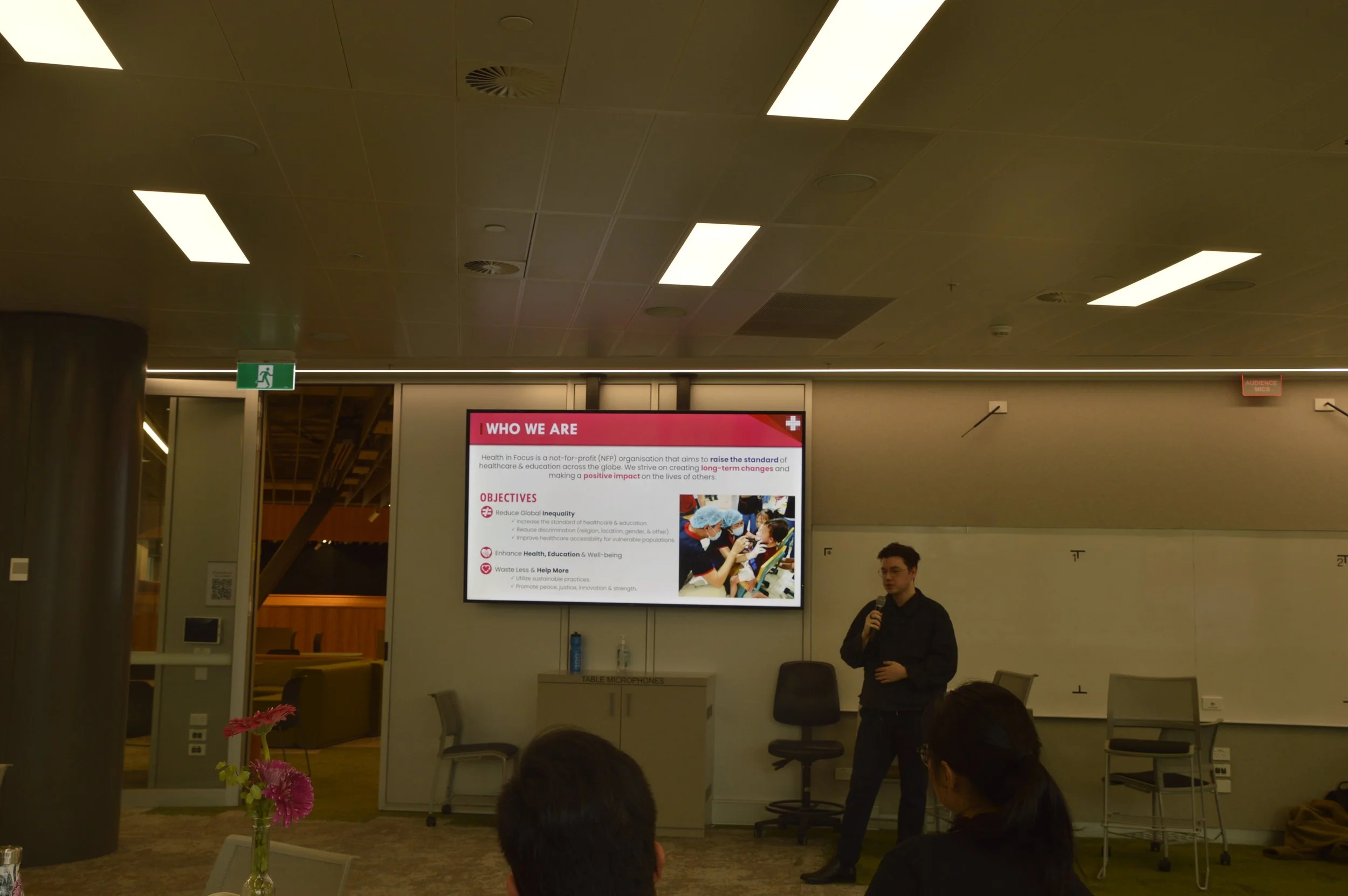 Speaker presenting a Health in Focus organization slide to an audience in a conference room.