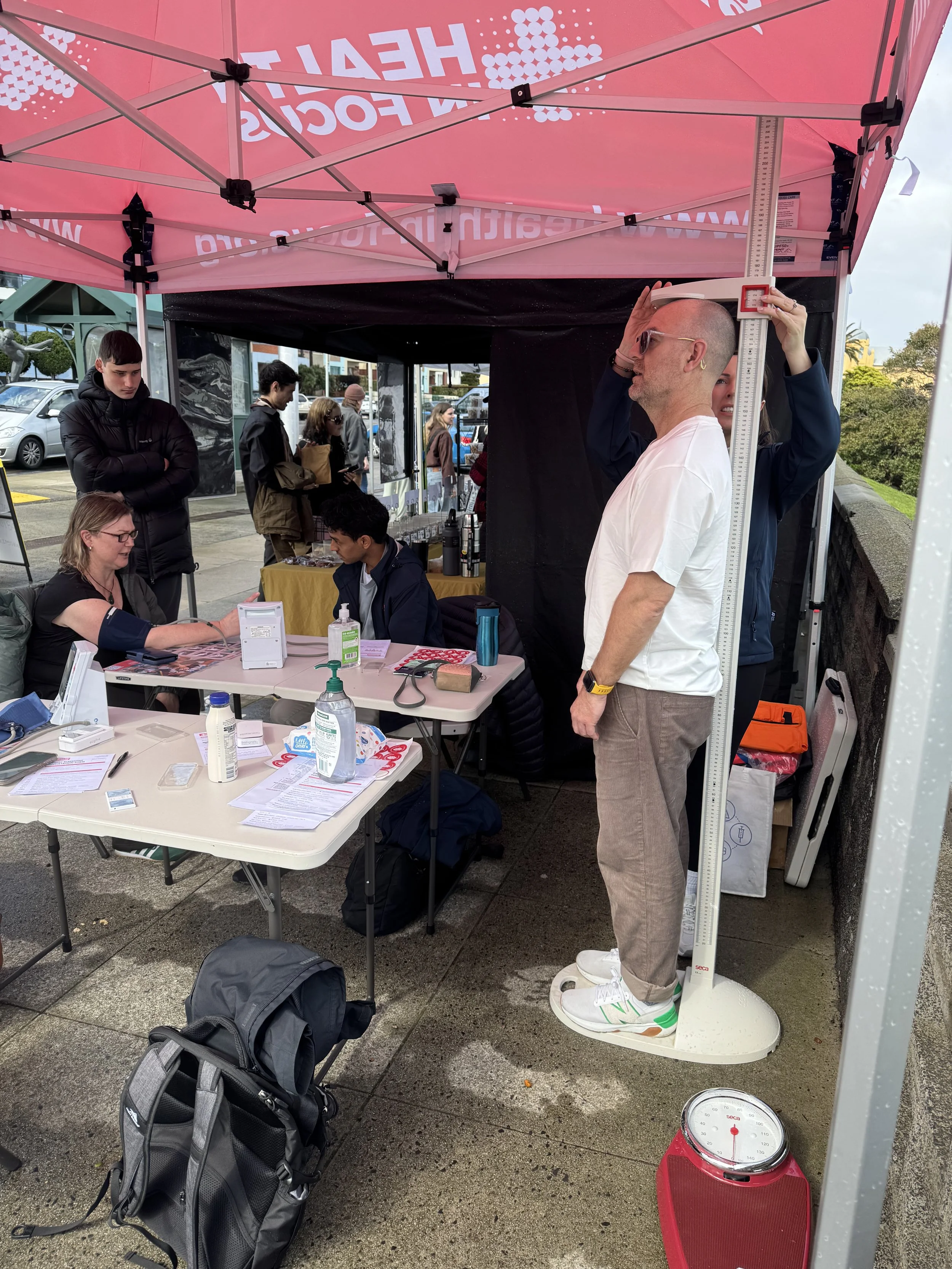 Community health screening under a pink tent, with staff measuring a man's height and checking participants' blood pressure and weight.