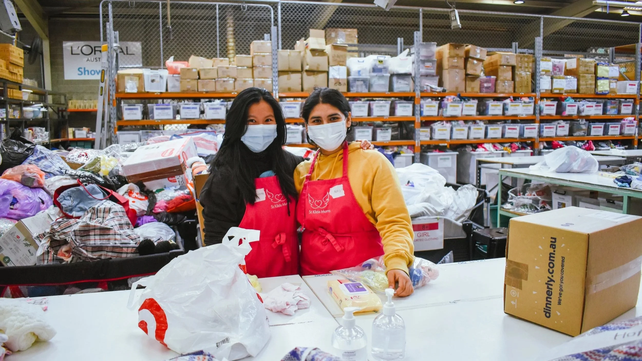 Two masked volunteers wearing red St Kilda Mums aprons sort donations in a clothing warehouse.