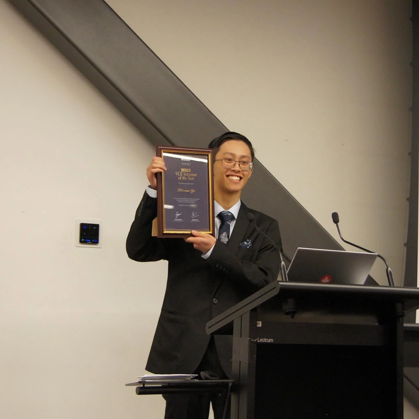 Man in a suit holding an award plaque beside a lectern and laptop