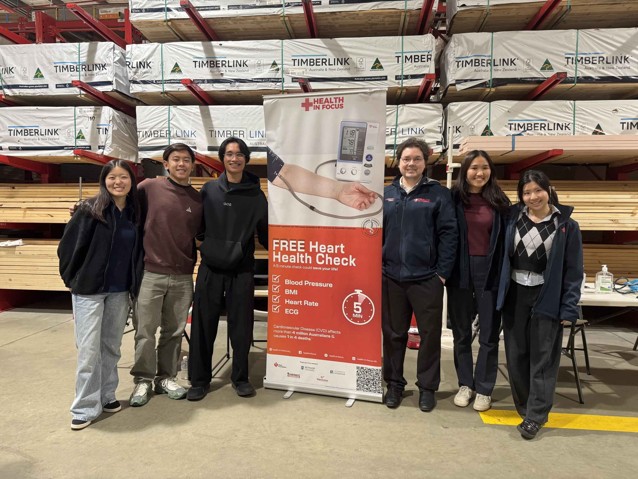 Six people pose beside a Health in Focus free heart health check banner in a timber warehouse.