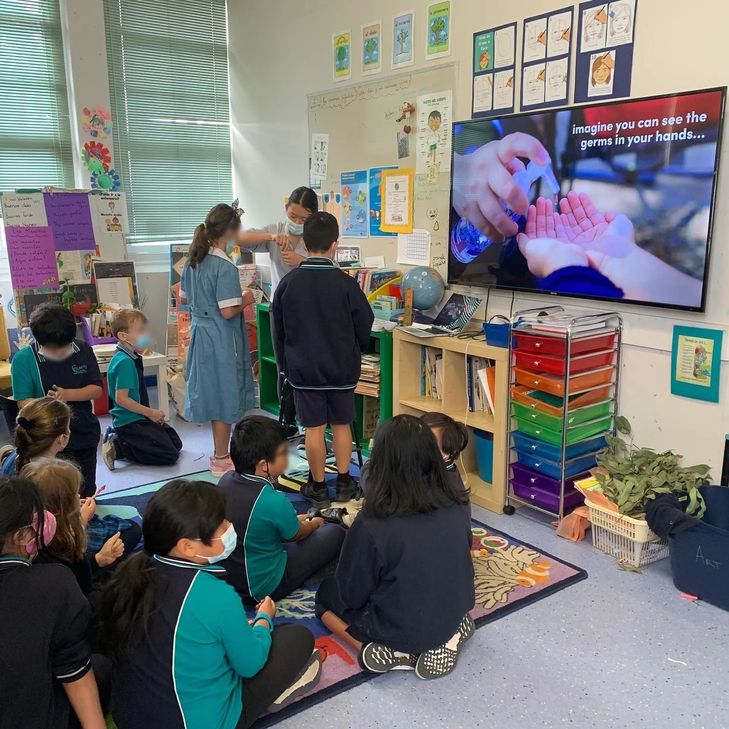 Elementary students watch a classroom handwashing lesson on a large screen while a teacher demonstrates using sanitizer.