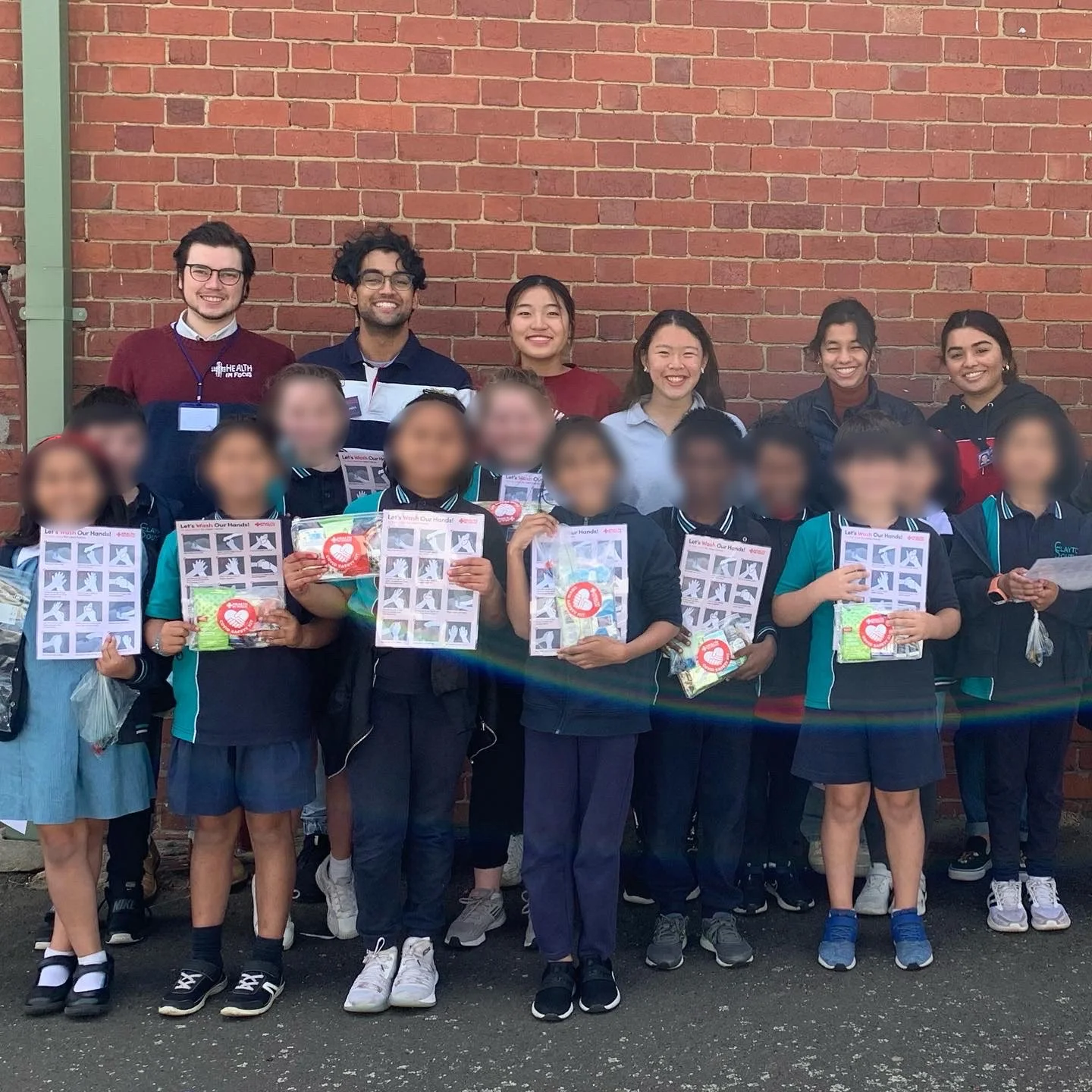 Group of students and volunteers holding handwashing education worksheets and hygiene supplies in front of a brick wall