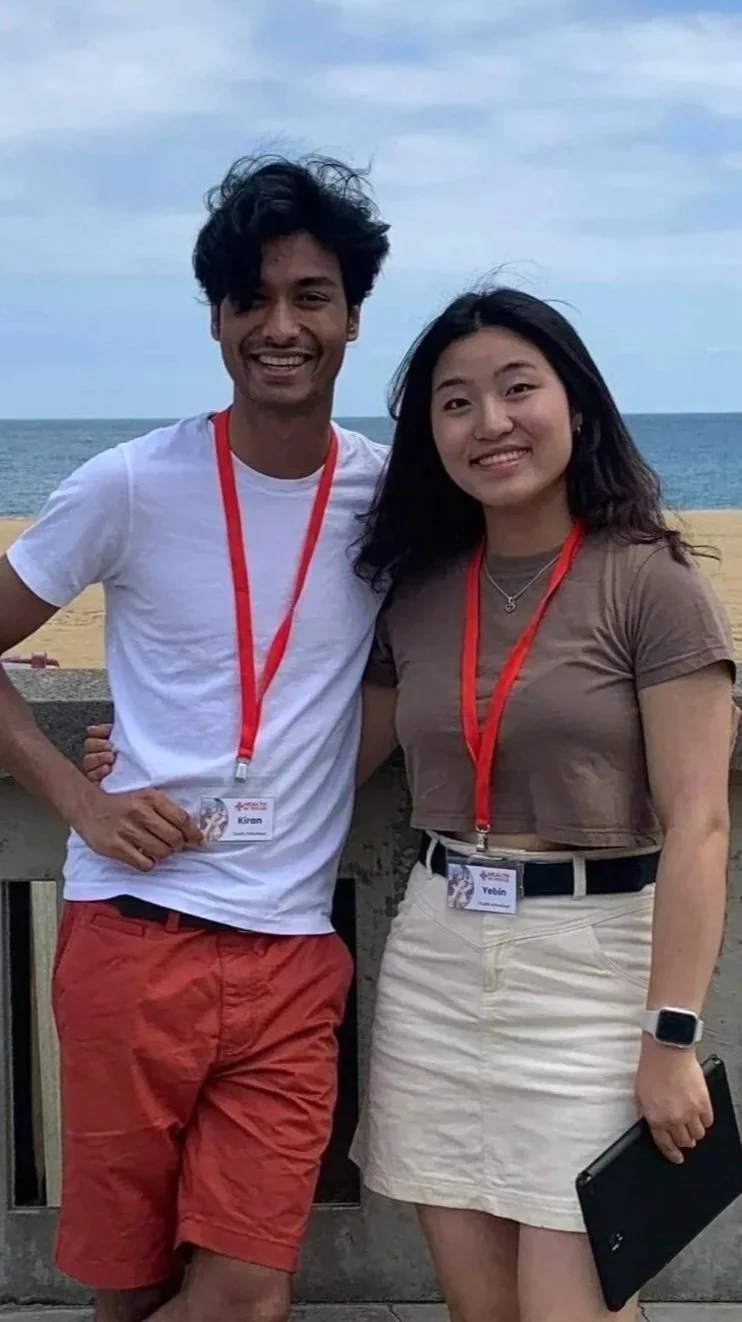Two smiling young adults wearing red event lanyards pose together on a beach with the ocean behind them.