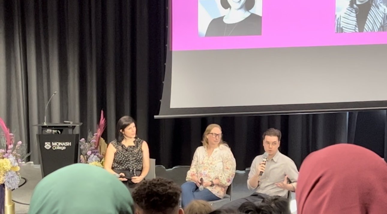 Panel discussion at Monash University with three speakers seated on stage, microphones in hand, and a presentation screen behind them.