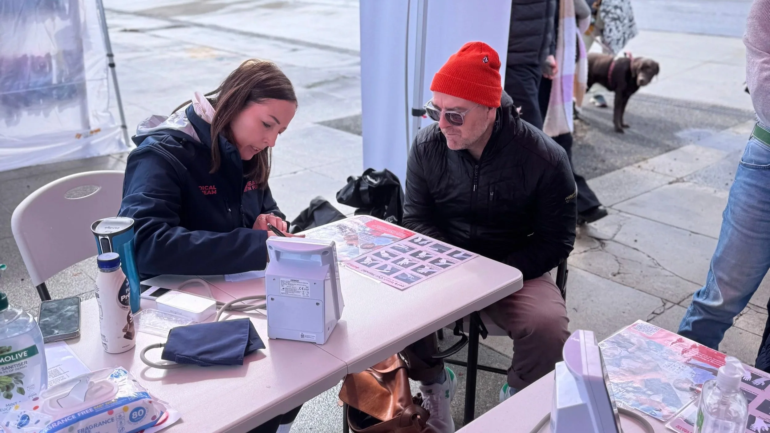 Woman in a medical volunteer jacket records information for a man wearing a red beanie at an outdoor health screening station.
