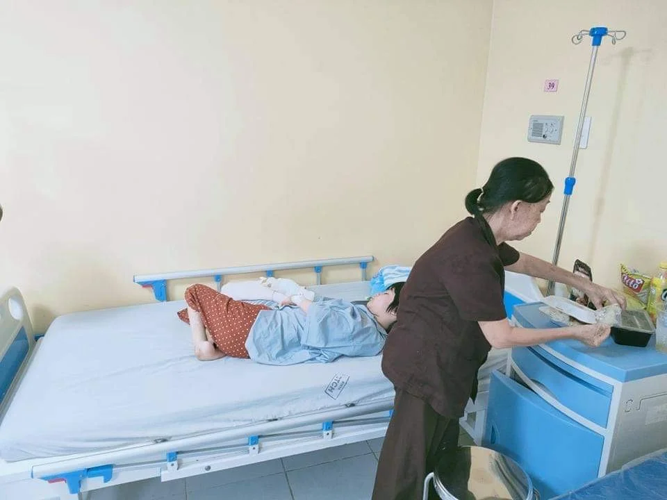 Hospital room with a patient resting in bed while a caregiver prepares supplies at a medical bedside cart.