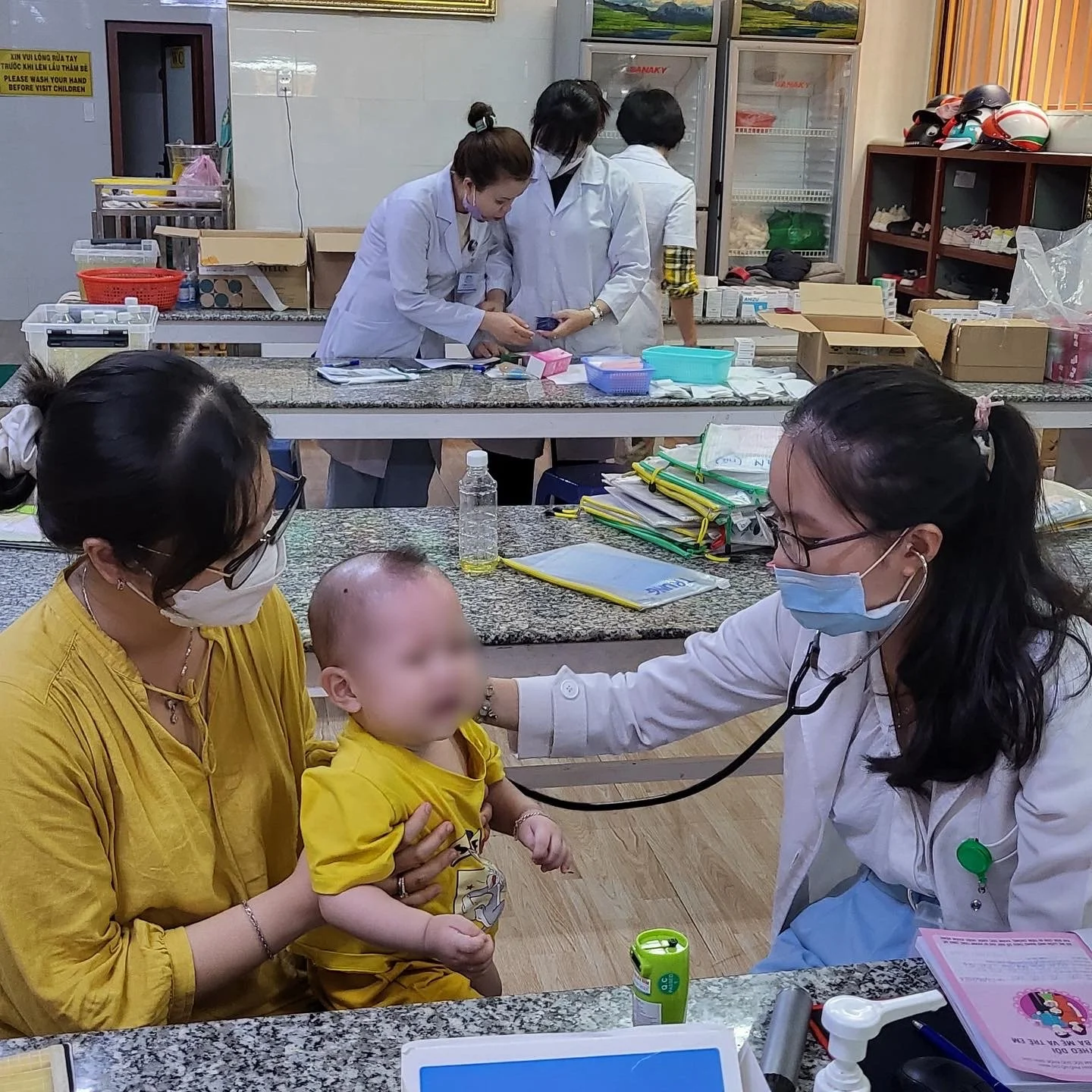Pediatrician examining a baby held by a masked mother in a clinic, with medical staff and supplies in the background