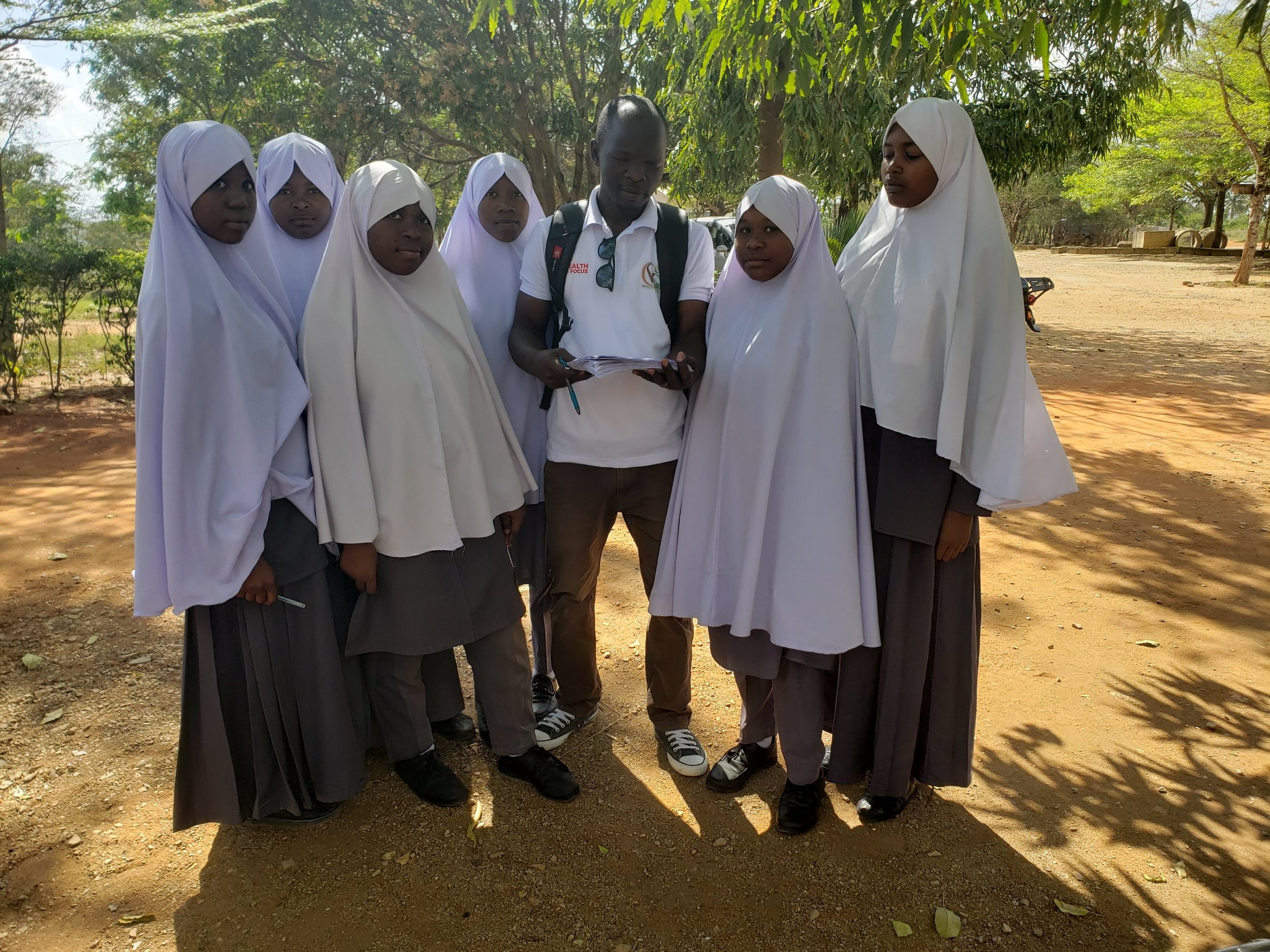 A man with a backpack stands with six schoolgirls wearing white hijabs and gray uniforms outdoors under trees.